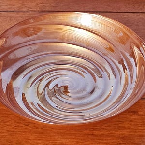 May include: A clear, oval-shaped glass bowl with a swirling pattern in shades of amber and white. The bowl's design creates a spiral effect, reflecting light and adding visual interest. The bowl is likely used for serving or display.