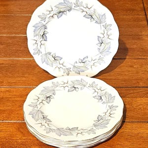 May include: A set of white ceramic plates with a delicate leaf design in shades of blue and gray. The plates have a scalloped edge and a gold trim. The plates are stacked on a wooden surface.