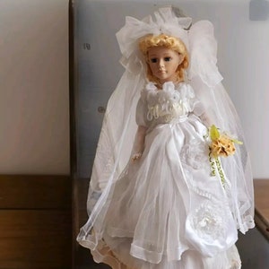 May include: A porcelain doll dressed in a white wedding gown with a veil and holding a bouquet of yellow flowers.