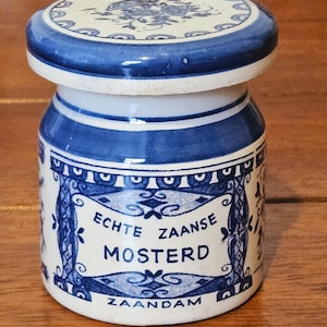 May include: A small, ceramic jar with a lid, featuring a blue and white floral design. The jar has a cylindrical shape with a blue rim and base. The text "Echte Zaanse Mosterd Zaandam" is displayed on the front.