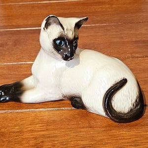 May include: A ceramic Siamese cat figurine in a reclining pose. The cat has a cream-colored body with dark brown points on the ears, face, paws, and tail. It features bright blue eyes and a glossy finish, capturing the classic color pattern.