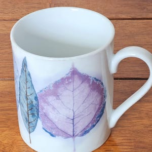 May include: White ceramic mug featuring a design of stylized leaves in shades of blue and purple. The mug has a curved handle and a smooth, glossy finish. The leaves are detailed with delicate veining.