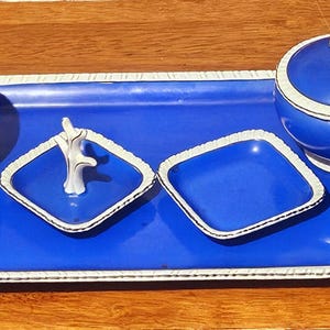 Könnte beinhalten: Ein kobaltblaues Servierset mit weißem Rand. Das Set beinhaltet ein rechteckiges Tablett, zwei Schalen mit Deckel und zwei quadratische Schalen. Eine Schale hat ein weißes Dekorationselement. Das Set steht auf einer Holzoberfläche.