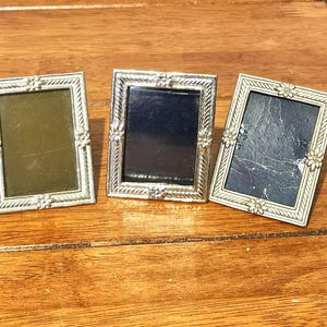 Könnte beinhalten: Drei kleine, rechteckige silberne Bilderrahmen mit floralen Eckakzenten. Jeder Rahmen hat eine unterschiedlich farbige Rückseite: Olivgrün, Schwarz und Dunkelblau. Die Rahmen sind auf einer Holzoberfläche angeordnet.