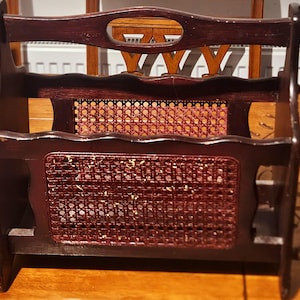 May include: A dark wood magazine rack with a woven cane design. The rack has two shelves and a handle at the top. The woven cane is a reddish-brown color, and the wood is a dark brown. The rack is designed to hold magazines or books.