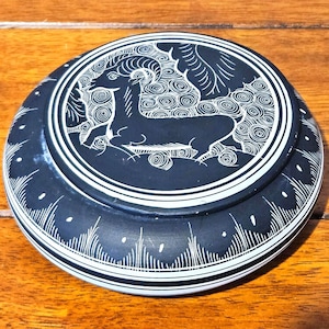 Puede incluir: Una caja redonda de piedra negra con una tapa que presenta un diseño blanco grabado de un carnero. La caja tiene un borde blanco y líneas blancas decorativas alrededor de los lados. La caja está sobre una superficie de madera.