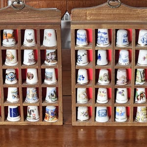 May include: Two wooden display cases, each with a grid of small compartments, showcasing a collection of decorative thimbles. The thimbles are white with various colourful designs, including floral patterns and scenic images. The cases have a dark wood finish.