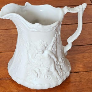 Puede incluir: Jarra de cerámica blanca con un diseño en relieve detallado. La jarra presenta un querubín y motivos florales. El asa está intrincadamente esculpida y el pico tiene un borde decorativo. Una pieza clásica para servir bebidas.