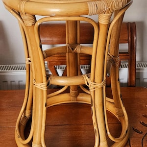 May include: A light brown rattan stool with a round woven seat. The stool has curved legs and is supported by a circular base. The frame is made of woven rattan and is tied together with woven rattan strips. The stool is on a wooden surface.