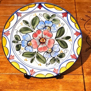 Op de afbeelding: Een decoratieve keramische plaat met een handgeschilderd bloemmotief. De plaat heeft een centrale opstelling van roze en blauwe bloemen met groene bladeren, omgeven door een rand van gele, blauwe en rode accenten. De plaat staat op een houten oppervlak.