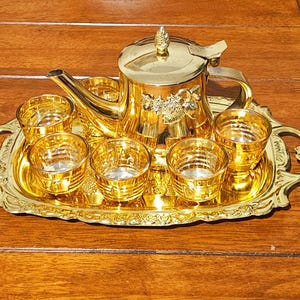 Könnte beinhalten: Goldfarbenes Teeservice mit Teekanne, sechs kleinen Gläsern und einem Tablett. Die Teekanne hat ein dekoratives Blumendesign und einen Deckel mit Knauf. Tablett und Gläser weisen aufwendige Details auf, was eine luxuriöse Ästhetik schafft.
