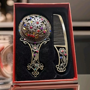 May include: A vintage vanity set comprising a hand mirror and comb. The bronze-toned mirror and comb are decorated with floral patterns and colourful gemstones. The set is presented in a red box with a black velvet interior.