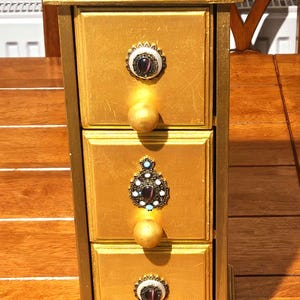 Può includere: Un portagioie in legno dipinto d'oro con tre cassetti. Ogni cassetto ha un pomello dorato e un accento decorativo color gioiello. La parte superiore della scatola è grigia. La scatola è su una superficie di legno.