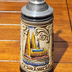 Peut inclure: Une chope à bière en céramique avec un relief détaillé de la cathédrale de Cologne, avec un couvercle et une poignée en argent. La chope est beige avec des accents colorés et les mots "Köln Dom" sont visibles.