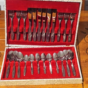 Puede incluir: Un juego de cubiertos vintage en una caja decorativa. El juego incluye tenedores, cuchillos y cucharas, todos con un acabado plateado brillante. El interior de la caja está forrado con terciopelo rojo, creando un contraste.