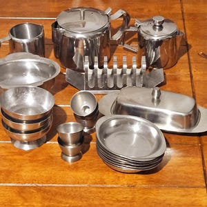 Puede incluir: Una colección de piezas de servicio de acero inoxidable, que incluye teteras, una jarra de crema, cuencos, una mantequera y una pila de pequeños platos ovalados. Los artículos están dispuestos sobre una superficie de madera.