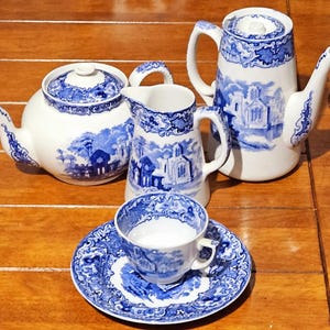 May include: A white and blue porcelain tea set featuring a teapot, creamer, sugar bowl, cup, and saucer. The set has a blue landscape design. The teapot has a curved spout and handle. The cup and saucer are small.