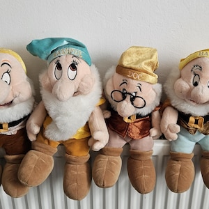 Op de afbeelding: Een set van vier pluche poppen die personages uit Sneeuwwitje en de zeven dwergen voorstellen. De poppen zijn gemaakt van zachte stof, hebben baarden van wit imitatiebont en gekleurde mutsen met geborduurde namen zoals Doc en Happy.