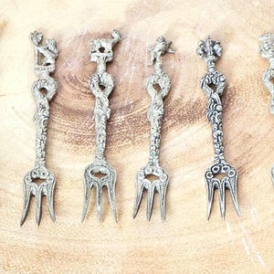 Può includere: Sei forchette antiche color argento con manici decorativi. Ogni forchetta presenta un design scolpito unico nella parte superiore, inclusi motivi floreali e animali. I rebbi sono sottili e le forchette sono disposte su una superficie di legno marrone chiaro.