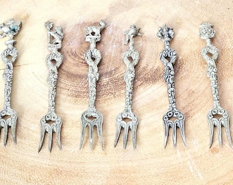 May include: Six antique silver-colored forks with decorative handles. Each fork features a unique sculpted design at the top, including floral and animal motifs. The tines are slender, and the forks are arranged on a light brown wooden surface.