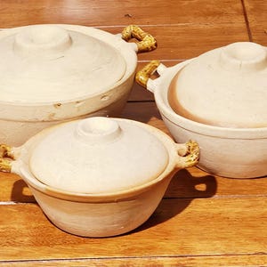 May include: Three beige ceramic cooking pots with lids, arranged on a wooden surface. The pots vary in size, each with a rounded shape and decorative handles. The lids have a central knob for easy lifting.