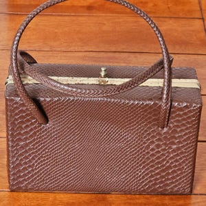 May include: A brown rectangular handbag with a snake-effect texture. It features a braided handle and a gold-coloured clasp. The top of the bag has gold-coloured trim. The handbag is on a wooden surface.