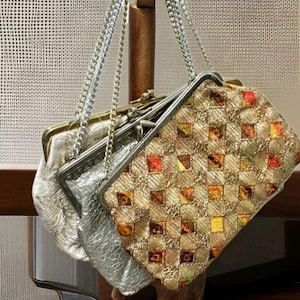 Puede incluir: Tres bolsos vintage con correas de cadena plateadas cuelgan de un soporte de madera. Los bolsos tienen marcos de metal plateado y dorado. Uno es plateado con un patrón floral en relieve, y otro es dorado con un patrón a cuadros.