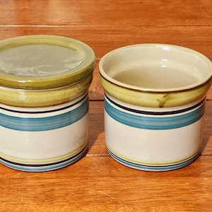 May include: Two ceramic jars with lids. The jars are white with a blue stripe, a black stripe, and a yellow-green rim. One jar has a lid, and the other is open. The jars are on a wooden surface.