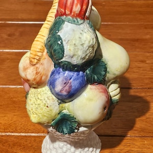 Può includere: Una scultura di un cesto di frutta in ceramica con una varietà di frutta colorata. La scultura comprende un carciofo con la parte superiore rossa, foglie verdi e una base a piedistallo bianca. I frutti sono dipinti in tonalità di giallo, blu e rosso.
