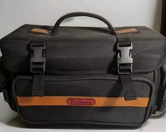 May include: Black camera bag with brown leather accents and a red "Pullman" logo.
