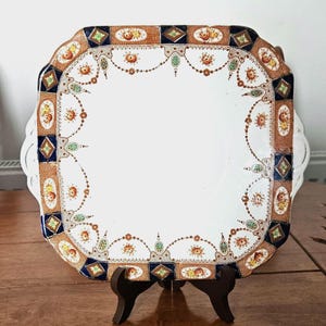 Puede incluir: Plato cuadrado de cerámica blanca con un borde decorativo. El borde presenta un patrón repetitivo de diseños florales y geométricos marrones, azul marino y dorados. El plato se exhibe sobre un soporte de madera marrón oscuro.