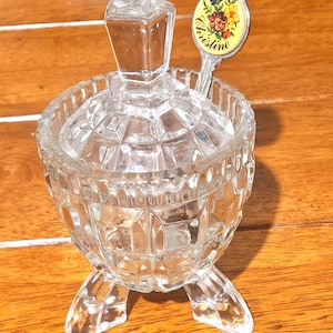 Könnte beinhalten: Eine klare, facettierte Zuckerdose aus Glas mit passendem Deckel und drei Füßen. Ein kleiner Löffel mit floralem Design und dem Namen "Christine" liegt in der Schüssel. Die Schüssel steht auf einer Holzoberfläche.