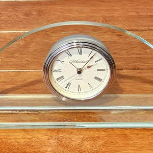 May include: A clear glass desk clock with a silver-tone clock face. The clock has Roman numerals and the brand name "Whitehall" is visible. The clock is set in a clear glass stand.