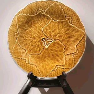 May include: A decorative plate with a leaf design. The plate features a central arrangement of large, detailed leaves in shades of brown and gold. The outer rim has a textured, cross-hatched pattern. The plate is displayed on a black stand.