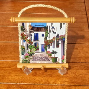 May include: A rectangular ceramic wall plaque featuring a colourful illustration of a street in Benalmádena, framed by light wood dowels and a thick rope hanger. The plaque includes three small wooden pegs at the bottom for hanging items.