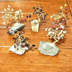 May include: Five gemstone trees with wire branches and stone bases are displayed on a wooden surface. The trees feature various colored gemstones, including clear, brown, and orange stones. The bases are rough-hewn rocks.