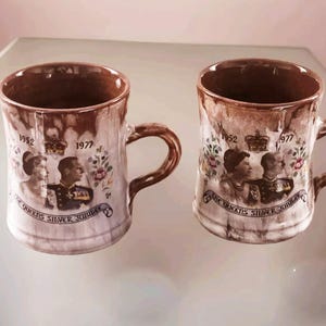 May include: Two brown ceramic mugs with a white glaze. The mugs feature a commemorative design of Queen Elizabeth II and Prince Philip with the text "The Queen's Silver Jubilee 1952 - 1977".