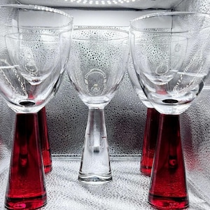 Könnte beinhalten: Vier klare Weingläser aus Glas mit roten Füßen. Die Gläser sind in einer Reihe auf einer silbernen Oberfläche angeordnet.