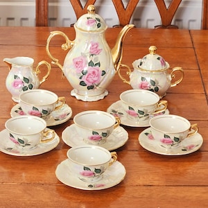 May include: A vintage tea set with a pearlescent finish, featuring pink rose floral designs. The set includes a teapot, sugar bowl, creamer, and multiple teacups with saucers. Gold accents adorn the handles and rims.