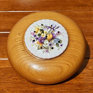 Könnte beinhalten: Ein runder Blumenfrosch aus Holz mit einem floralen Keramikeinsatz. Der Einsatz zeigt einen Strauß aus lila, gelben und blauen Blumen. Der Holzsockel ist hellbraun und hat eine glatte Oberfläche.