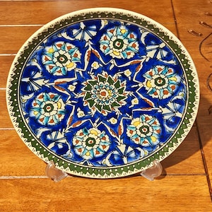 Puede incluir: Un plato decorativo de cerámica con un vibrante fondo azul e intrincados diseños florales en tonos verdes, amarillos y blancos. El plato tiene un borde blanco y un borde verde con un diseño punteado. El plato se exhibe sobre un soporte transparente.