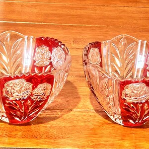 May include: Two clear crystal bowls with a red floral design. The bowls have a heart-shaped design with a scalloped edge. The floral design includes roses and leaves. The bowls are on a wooden surface.