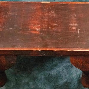 May include: A small, rectangular wooden step stool with a dark brown finish. The stool has a slightly distressed appearance, with visible wear and tear on the surface. It features four curved legs, adding a touch of elegance to its design. The stool measures approximately 12 inches long, 8 inches wide, and 6 inches high.
