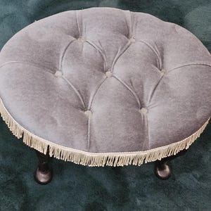 Puede incluir: Un otomán gris de forma ovalada con la parte superior acolchada y ribete de flecos. El otomán tiene cuatro patas oscuras y curvadas. La parte superior acolchada tiene un diseño de botones. El ribete de flecos es de color crema claro.