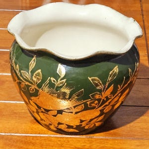 Op de afbeelding: Een decoratieve keramische plantenbak met een donkergroene buitenkant en een witte binnenkant. De plantenbak heeft gouden blad- en takontwerpen. De rand heeft een geschulpte rand.