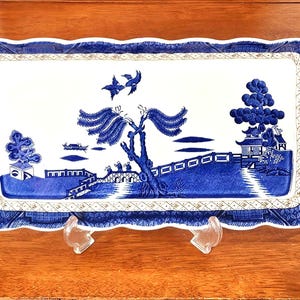 Peut inclure: Un plateau de service rectangulaire en céramique avec un motif bleu et blanc. Le décor représente un paysage avec un pont, un grand arbre, deux oiseaux en vol et des pagodes. Le plateau présente un bord festonné et une bordure décorative.