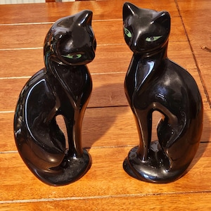 May include: Two glossy black cat figurines with green eyes sit side-by-side. The cats are in a seated position, with their tails curled around their bodies. The figurines are on a wooden surface.