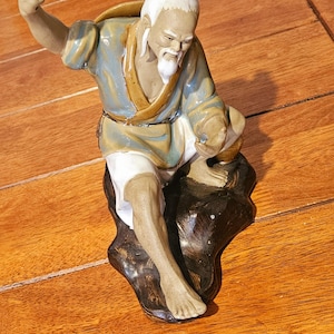 May include: A ceramic figurine of an elderly person with a white beard and hair, wearing a blue and tan robe over white pants. The figure is posed in a seated position on a dark brown rock-like base with one arm raised and a fist clenched.