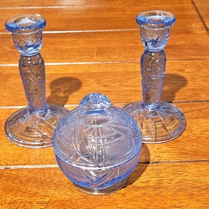 May include: A set of three blue glass decorative items. Two matching candle holders with detailed stems and circular bases flank a small, lidded jar. The glass has a textured, cut-glass appearance, reflecting light. The items are arranged on a wooden surface.