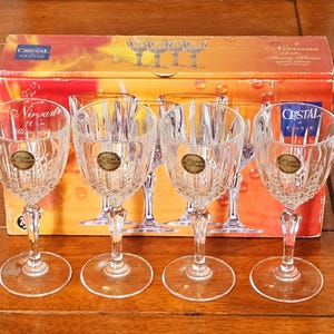 Puede incluir: Juego de seis copas de jerez de cristal transparente con diseño acanalado, expuestas frente a una caja naranja y dorada. La caja presenta la marca "Cristal" y las palabras "Nirvana" y "Sherry Glasses".